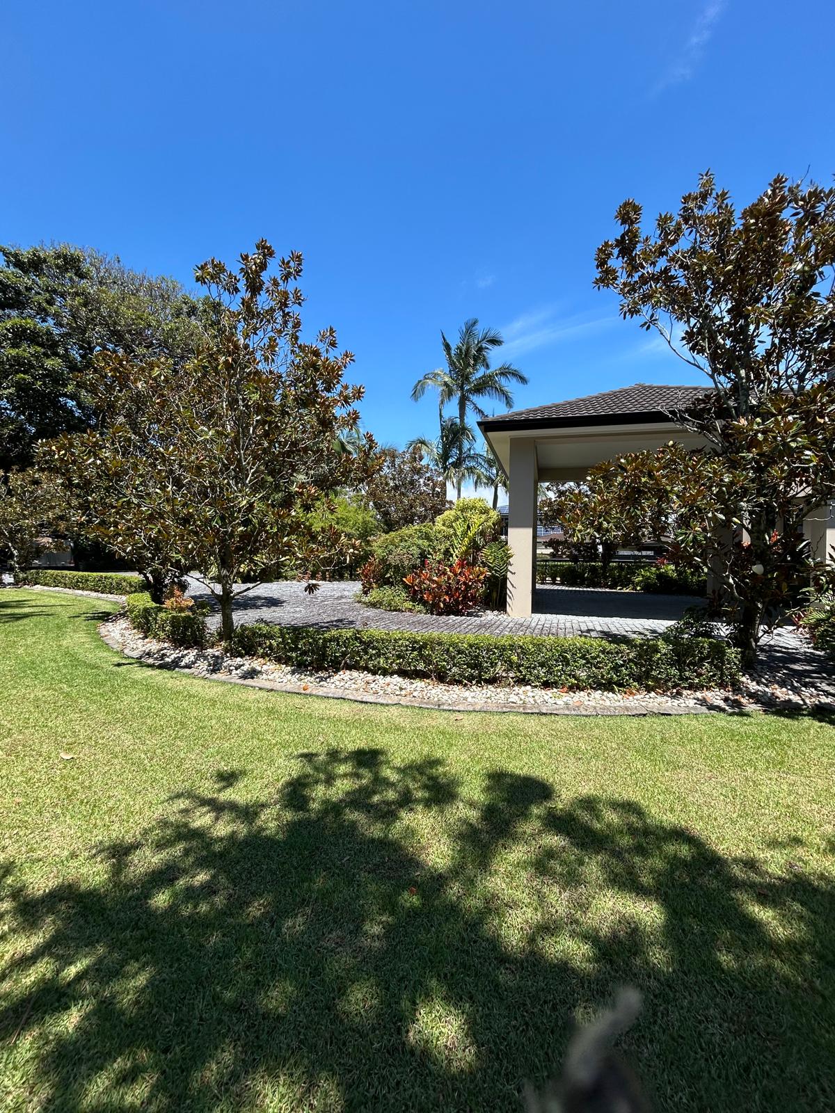 Sweeping green lawn with magnolia trees, trimmed hedges, decorative stone borders, and a covered portico at a prestige home