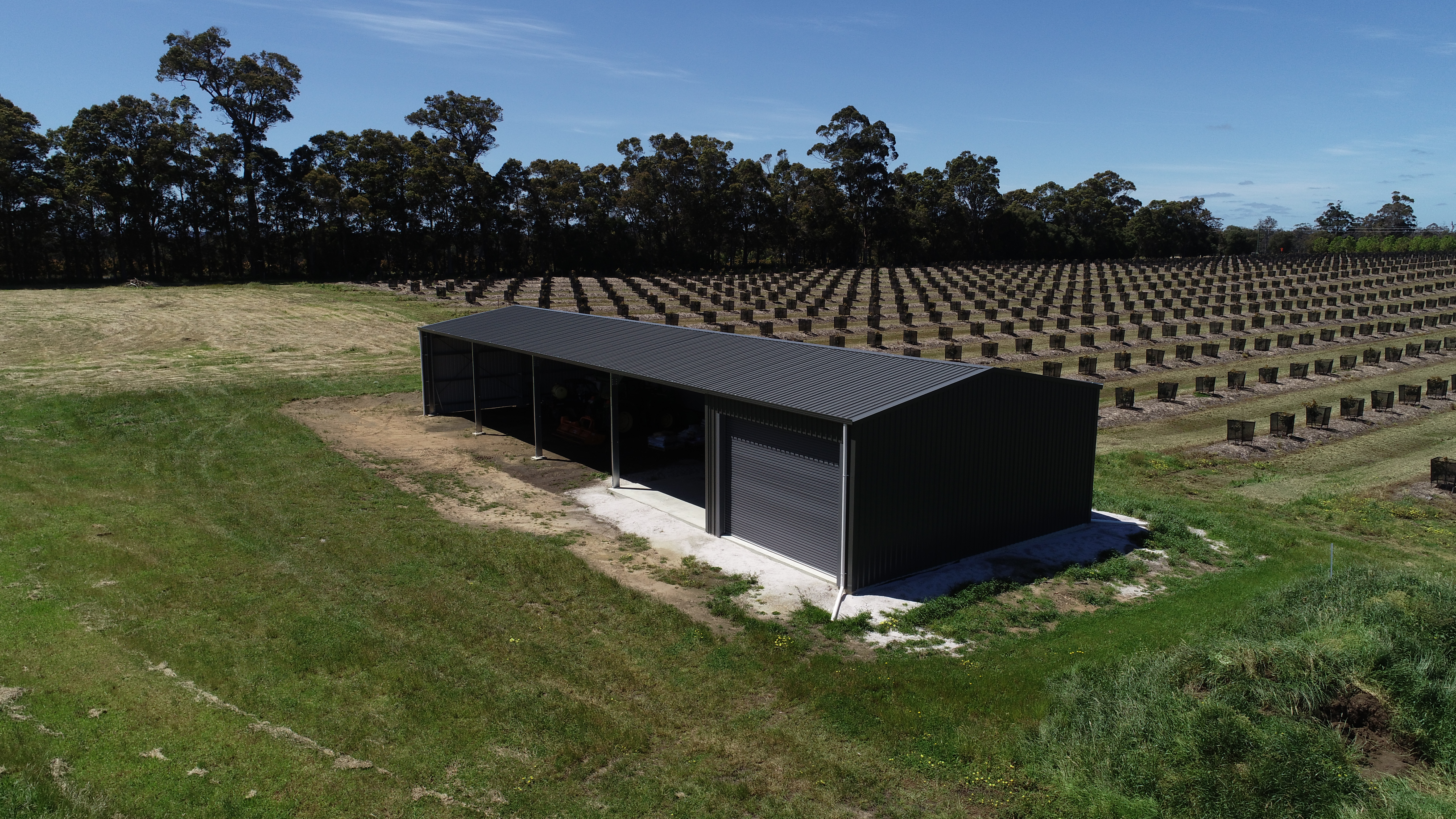 Dark steel farm shed with open bays and a roller door beside an orchard