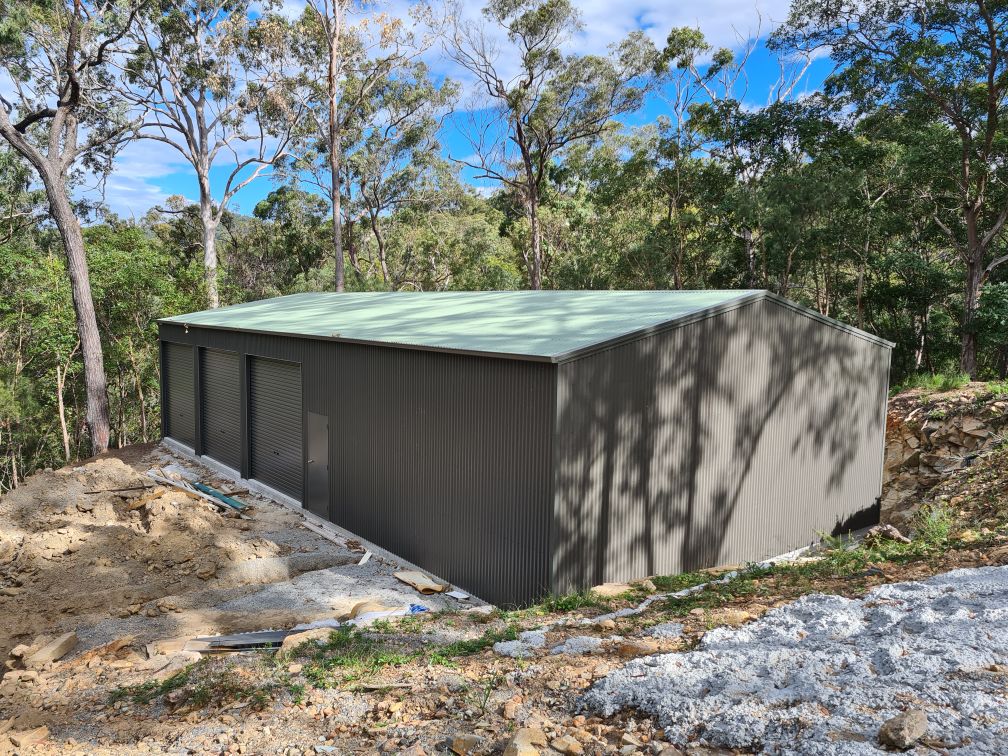 Dark steel building with three roller doors and a green roof cut into a bush slope
