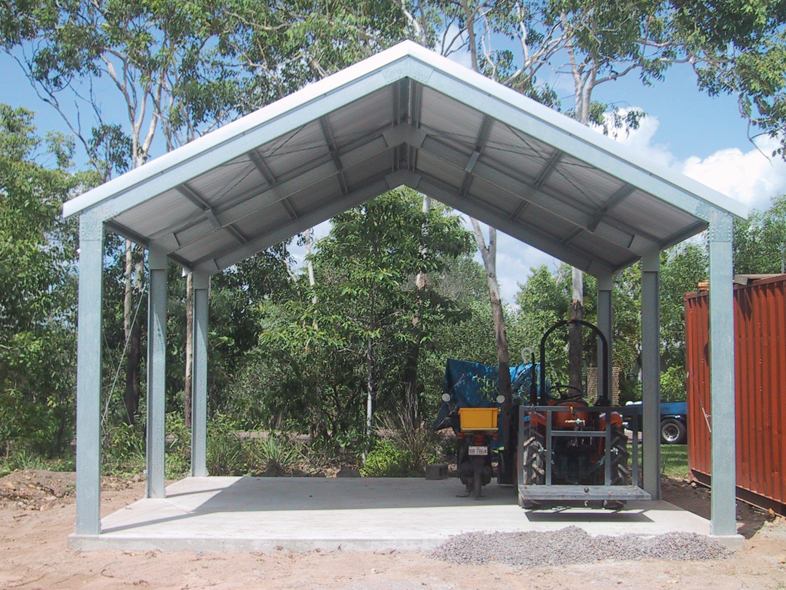 Open steel gable carport with a tall roof pitch and a tractor parked underneath